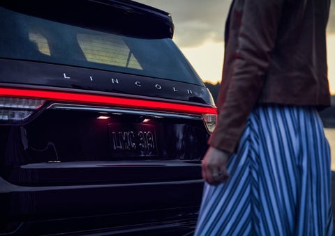 A person is shown near the rear of a 2024 Lincoln Aviator® SUV as the Lincoln Embrace illuminates the rear lights | Angela Krause Lincoln of Alpharetta in Alpharetta GA